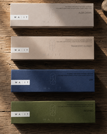 Four rectangular product boxes arranged vertically on a rustic wooden surface, each in a different color: beige, light beige, navy blue, and olive green. The boxes are branded WA:IT and labeled with scent names including Alba Dawn, Tramonto Sunset, UMI, and Respiro.